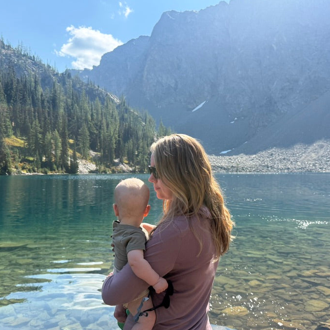 Woman holding a baby by a lake with mountains in the background